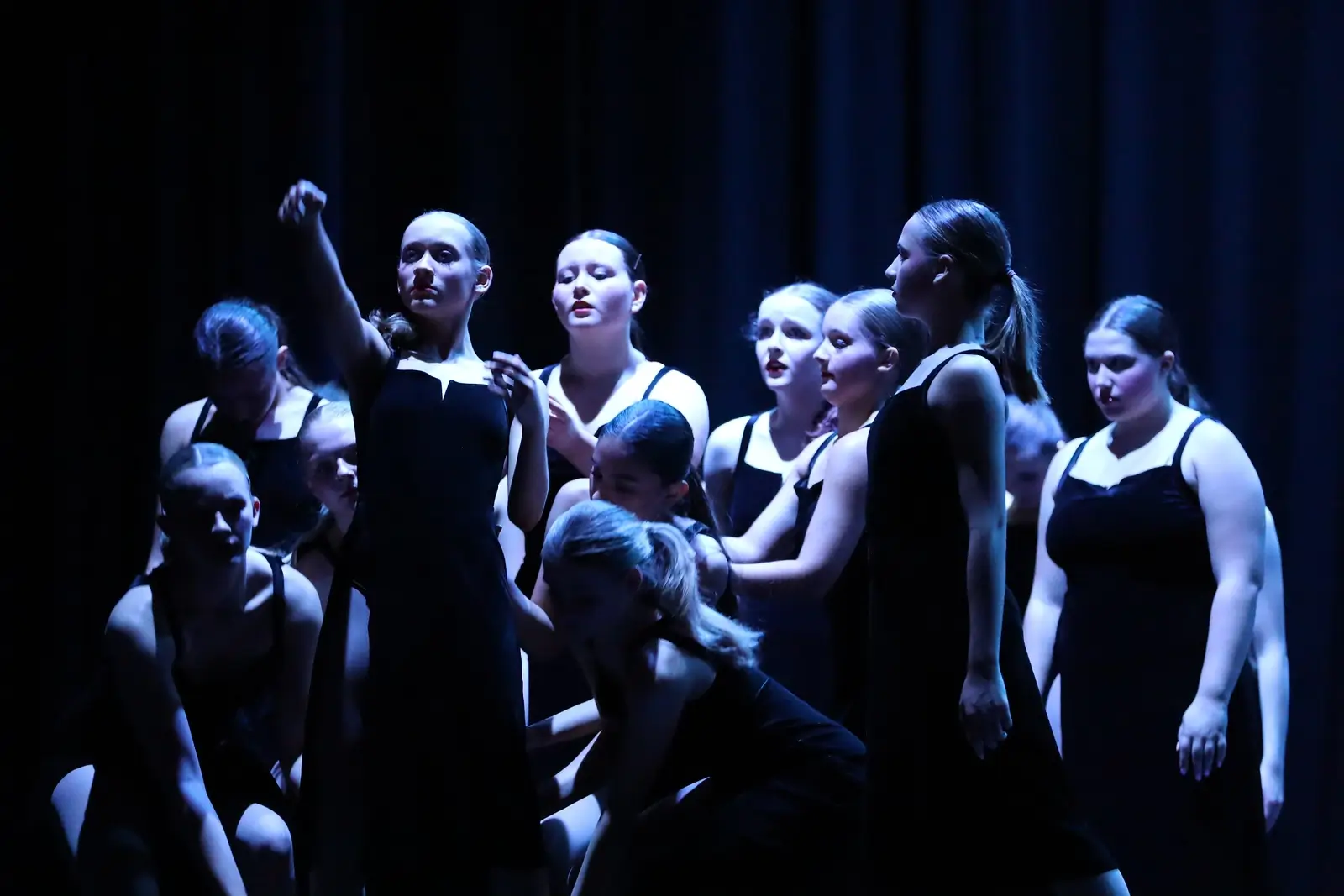 A group of dancers in black dresses perform on a dimly lit stage, with one dancer in front pointing outward and others gathered closely behind her, all under dramatic blue lighting—a striking showcase of ballet tap jazz lessons. -Showstoppers Dance Studio