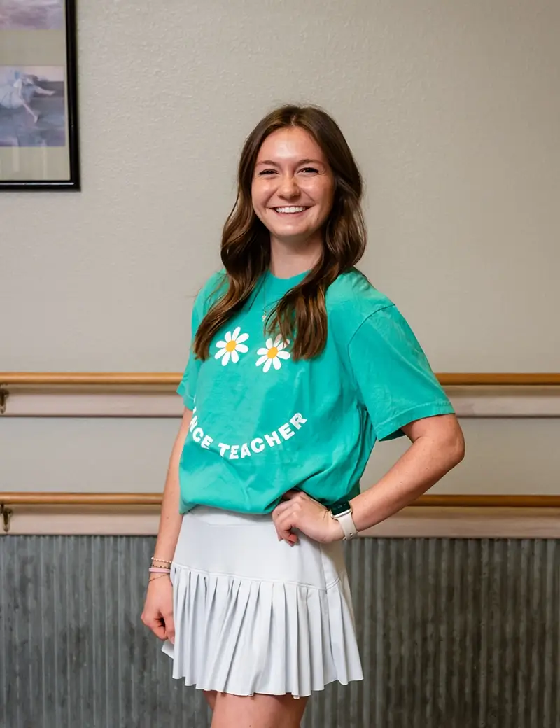A smiling woman with long brown hair, wearing a green "DANCE TEACHER" t-shirt and a white pleated skirt, stands in a dance studio offering ballet tap jazz lessons, perfect for kids dance classes or Dance lessons Stillwater. -Showstoppers Dance Studio