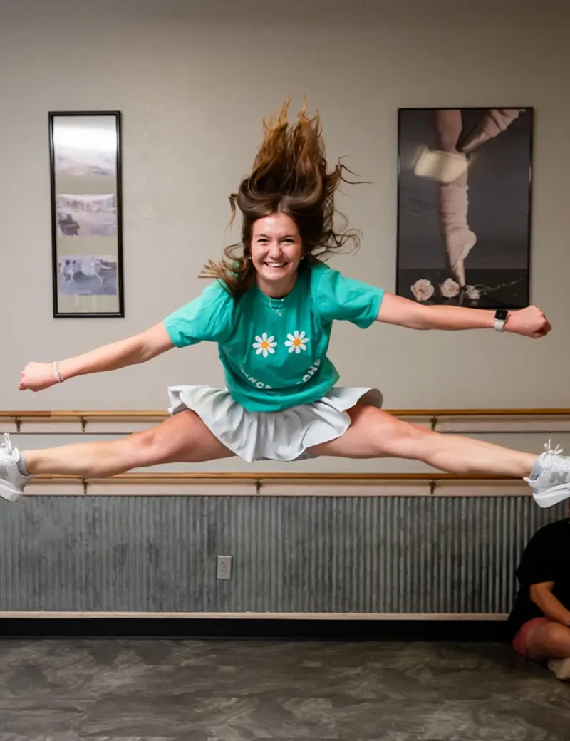 A young woman in a green shirt and white skirt performs a mid-air split jump in a dance studio, smiling. Her hair is flying up, and ballet tap jazz lessons posters are on the wall behind her. -Showstoppers Dance Studio