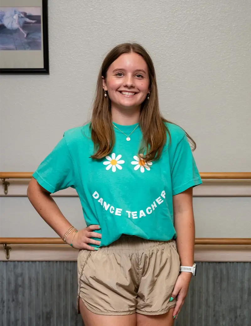 A young woman with long brown hair smiles at the camera, wearing a green Dance Teacher t-shirt with flower graphics and beige shorts. She stands indoors at a dance studio, in front of a ballet barre and a light-colored wall. -Showstoppers Dance Studio