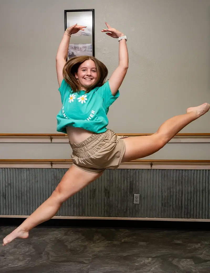 A young woman in a green daisy t-shirt and tan shorts leaps joyfully in a dance studio during adult dance classes in Stillwater, smiling and extending her arms and legs, with her hair flying mid-jump. -Showstoppers Dance Studio