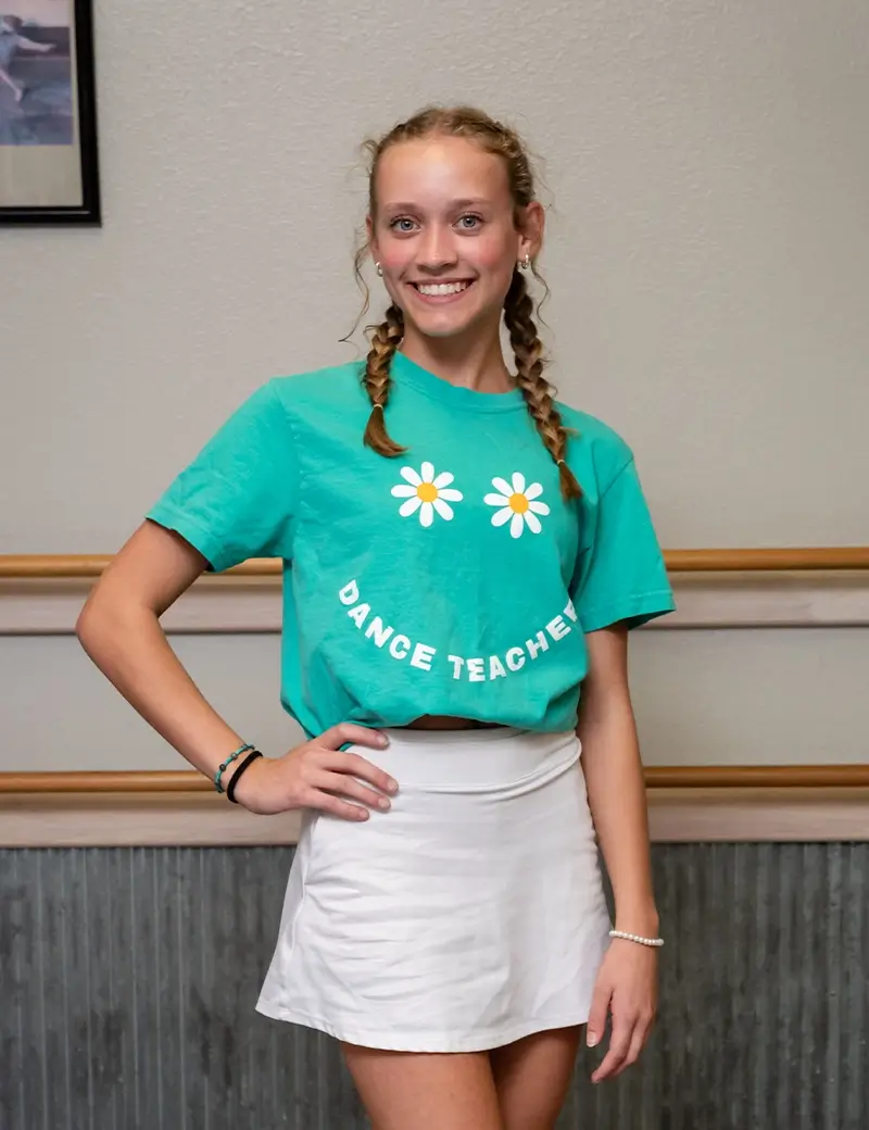 A young woman with braided hair smiles, wearing a green Dance Teacher t-shirt with daisy designs and a white skirt, standing indoors against a beige wall at a welcoming dance studio. -Showstoppers Dance Studio