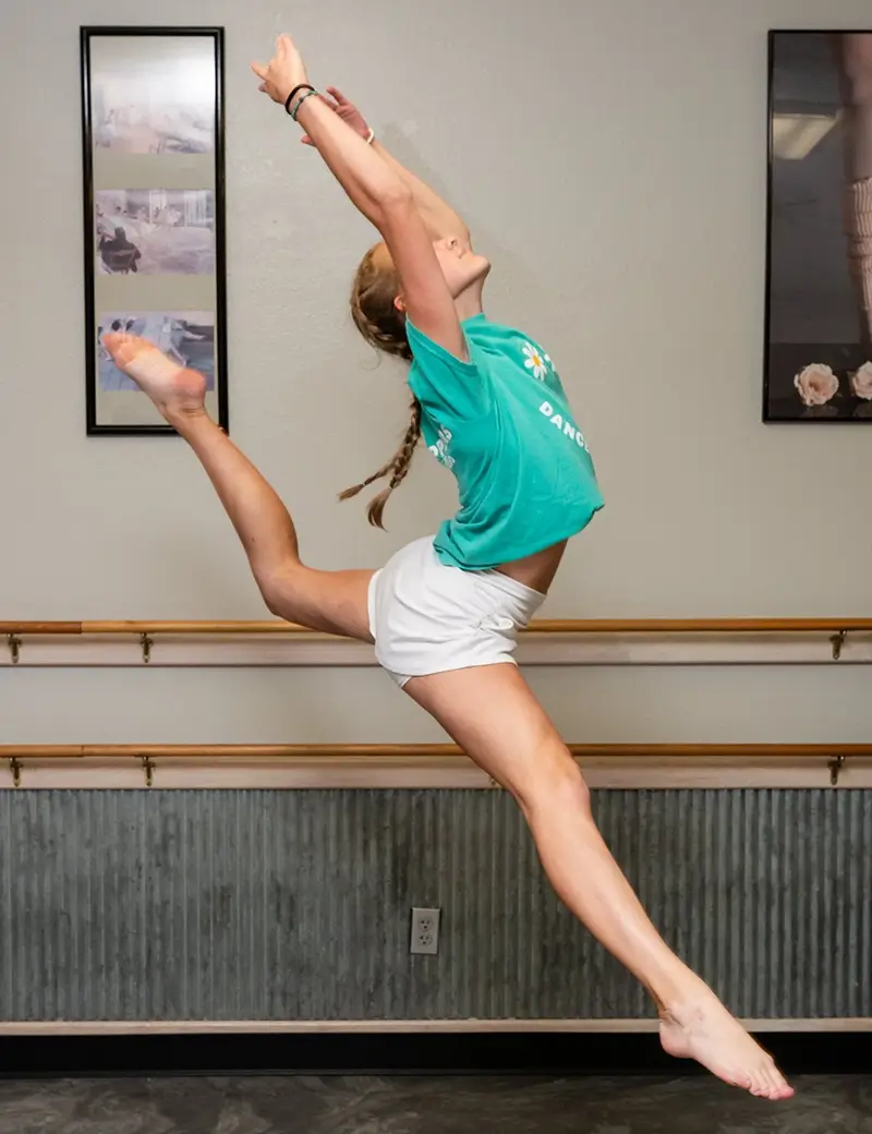 A girl in a green T-shirt and white shorts leaps gracefully in a dance studio, with one arm and one leg extended upward. Ballet barres and framed dance photos line the wall, capturing the joy of kids dance classes. -Showstoppers Dance Studio