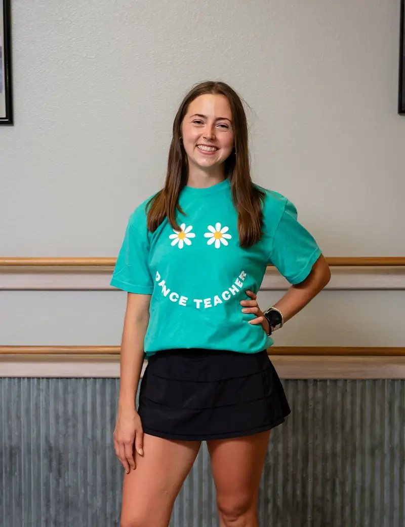 A smiling young woman with long brown hair stands indoors at a dance studio, wearing a teal Dance Teacher T-shirt with daisy graphics and a black skirt. She has one hand on her hip and is posing against a neutral wall. -Showstoppers Dance Studio