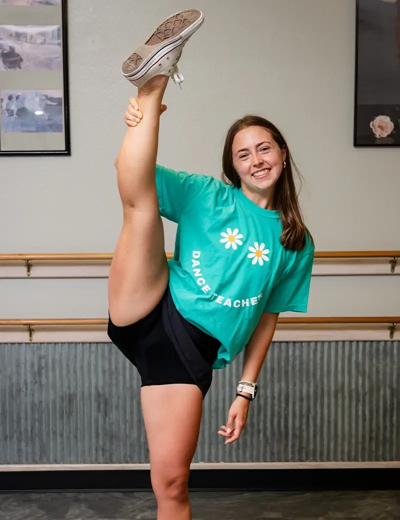 A smiling woman in a green Dance Teacher t-shirt and black shorts stands in a dance studio, holding one leg straight up high beside her head. Framed photos and a ballet barre highlight the inspiring space for adult dance classes. -Showstoppers Dance Studio