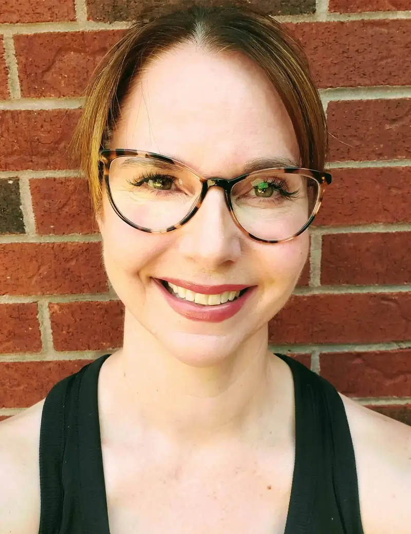 A woman with light skin, brown hair pulled back, and wearing tortoiseshell glasses smiles at the camera. She is dressed in a black top and stands in front of a red brick wall at a vibrant dance studio offering ballet, tap, and jazz lessons. -Showstoppers Dance Studio