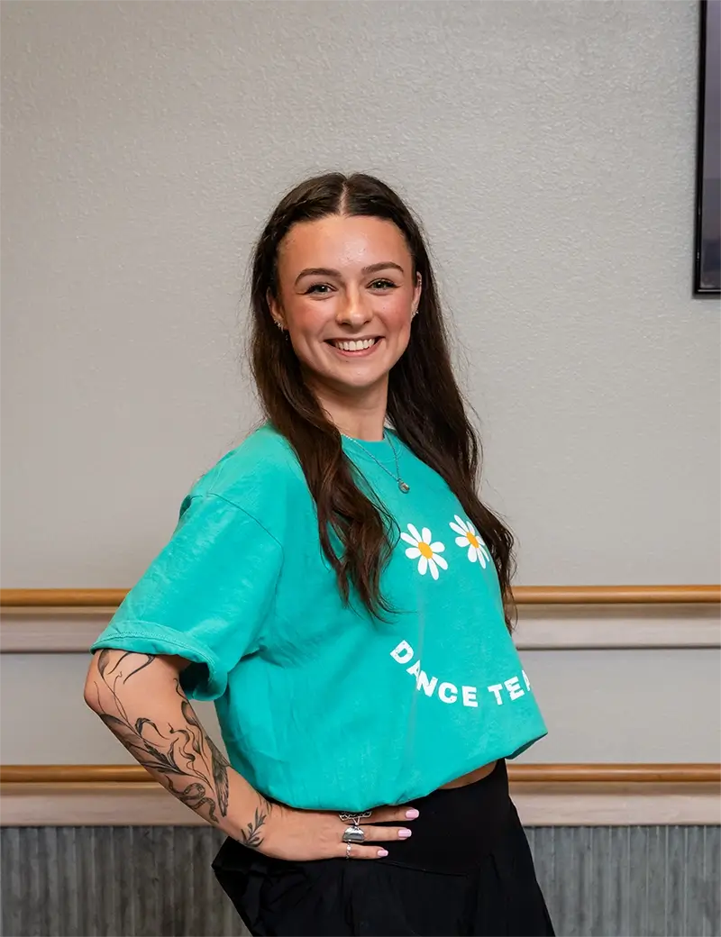 A woman with long brown hair smiles at the camera. Wearing a teal DANCE TEAM T-shirt, she stands in front of a neutral wall—ready for ballet tap jazz lessons at her favorite dance studio in Stillwater. A floral tattoo decorates her left arm. -Showstoppers Dance Studio