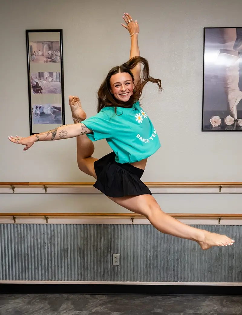 A young woman in a turquoise T-shirt and black shorts performs an energetic leap in a dance studio, smiling with arms outstretched. Art and dance posters hang on the wall behind her—perfect inspiration for Dance classes Stillwater. -Showstoppers Dance Studio