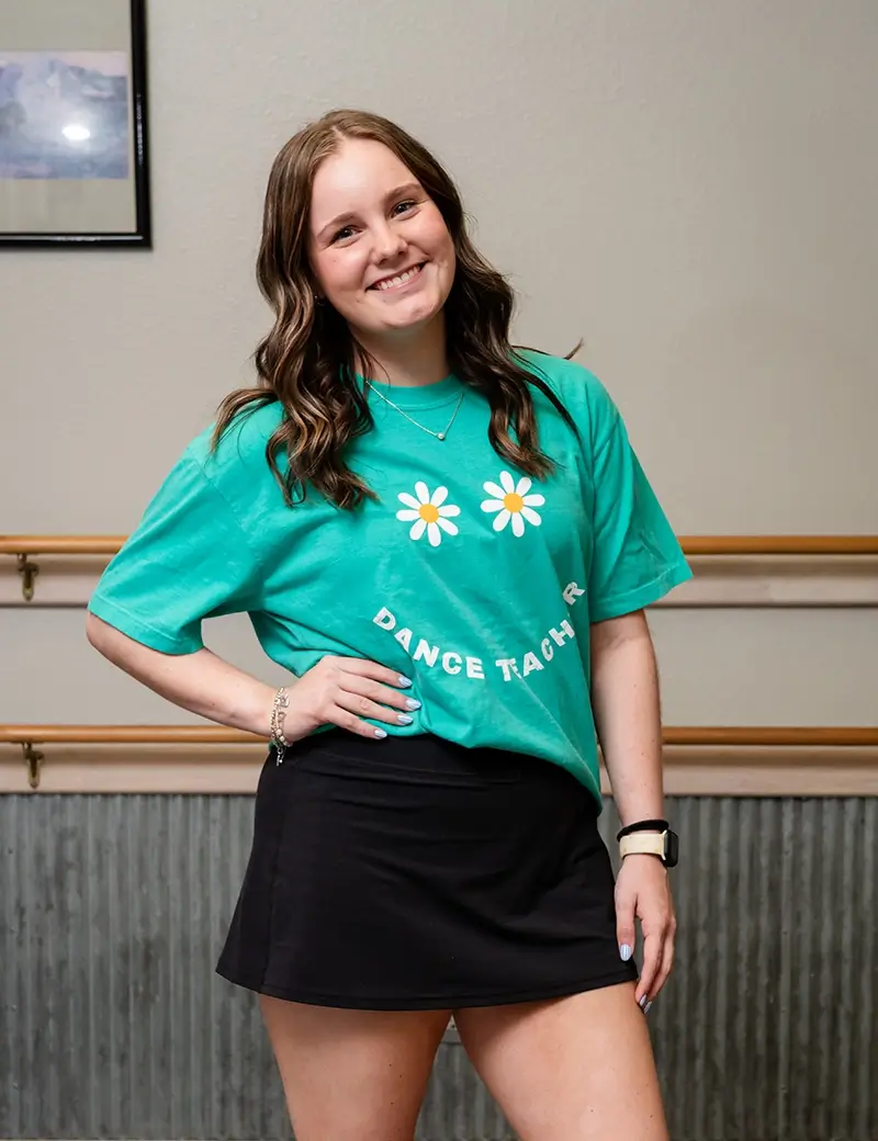 A smiling woman with wavy brown hair stands indoors wearing a green Dance Teacher t-shirt with daisy graphics and a black skirt. She poses confidently in a dance studio, ready to lead kids dance classes or dance lessons Stillwater families love. -Showstoppers Dance Studio