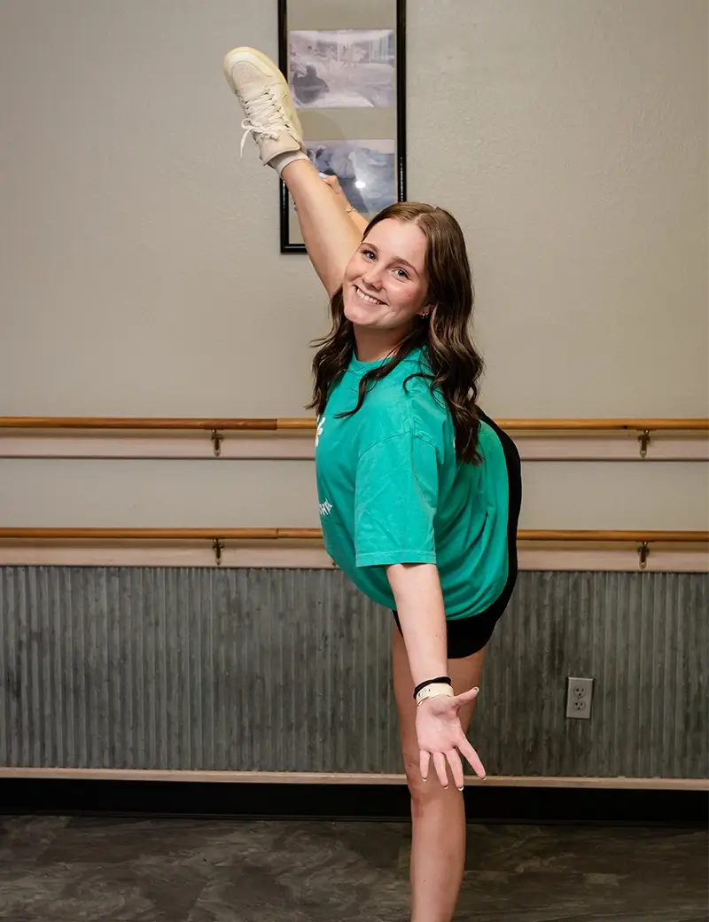 A young woman in a green shirt and black shorts stands on one leg in a dance studio, smiling at the camera while lifting her other leg high above her head during dance lessons in Stillwater. -Showstoppers Dance Studio