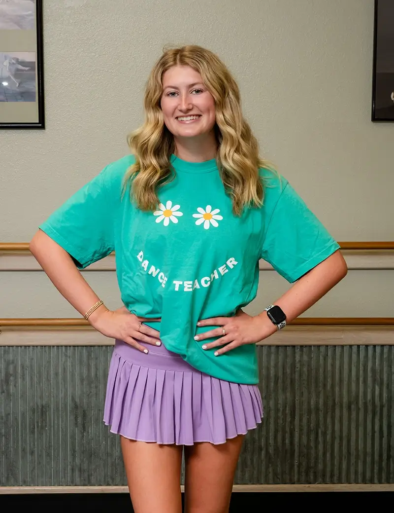 A smiling young woman with wavy blonde hair wears a green Dance Teacher t-shirt with daisies and a pleated lavender skirt, standing with hands on hips in a dance studio where kids dance classes like ballet, tap, and jazz lessons are offered. -Showstoppers Dance Studio