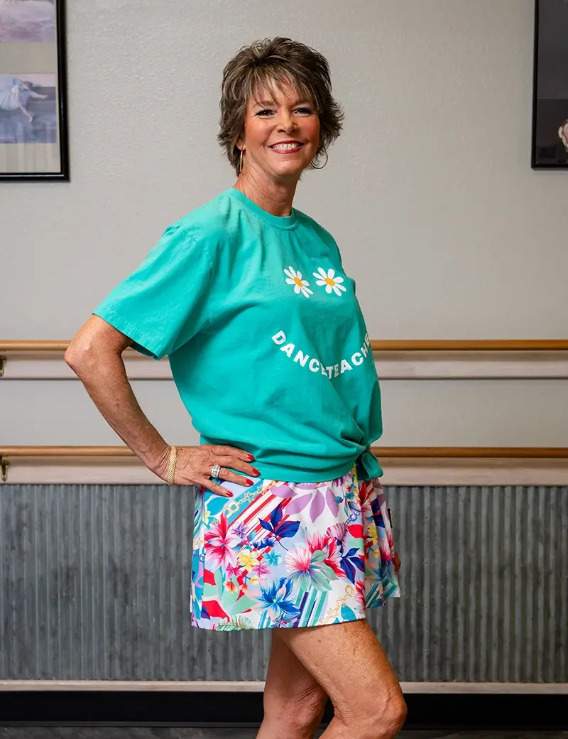 A smiling woman stands indoors, wearing a teal Dance Teacher T-shirt with daisy graphics and colorful floral shorts. She poses in a dance studio, ready to inspire young dancers in ballet, tap, and jazz lessons at Kids dance classes. -Showstoppers Dance Studio