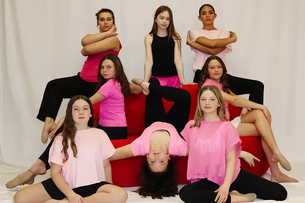 Eight girls pose in dance attire on and around a red couch against a white backdrop. Some wear pink tops, others black. One girl is upside down in a backbend in front, while others stand or sit confidently. -Showstoppers Dance Studio