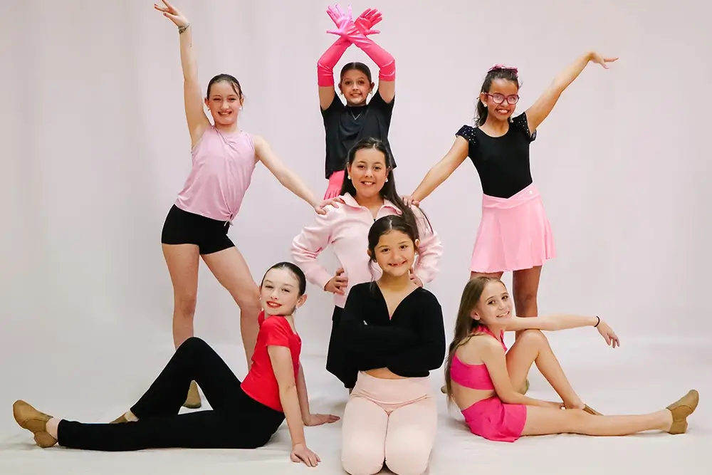 Seven young girls in colorful dance outfits pose cheerfully against a plain white background. Some are standing with arms up, while others are sitting or kneeling, all smiling and looking at the camera. -Showstoppers Dance Studio