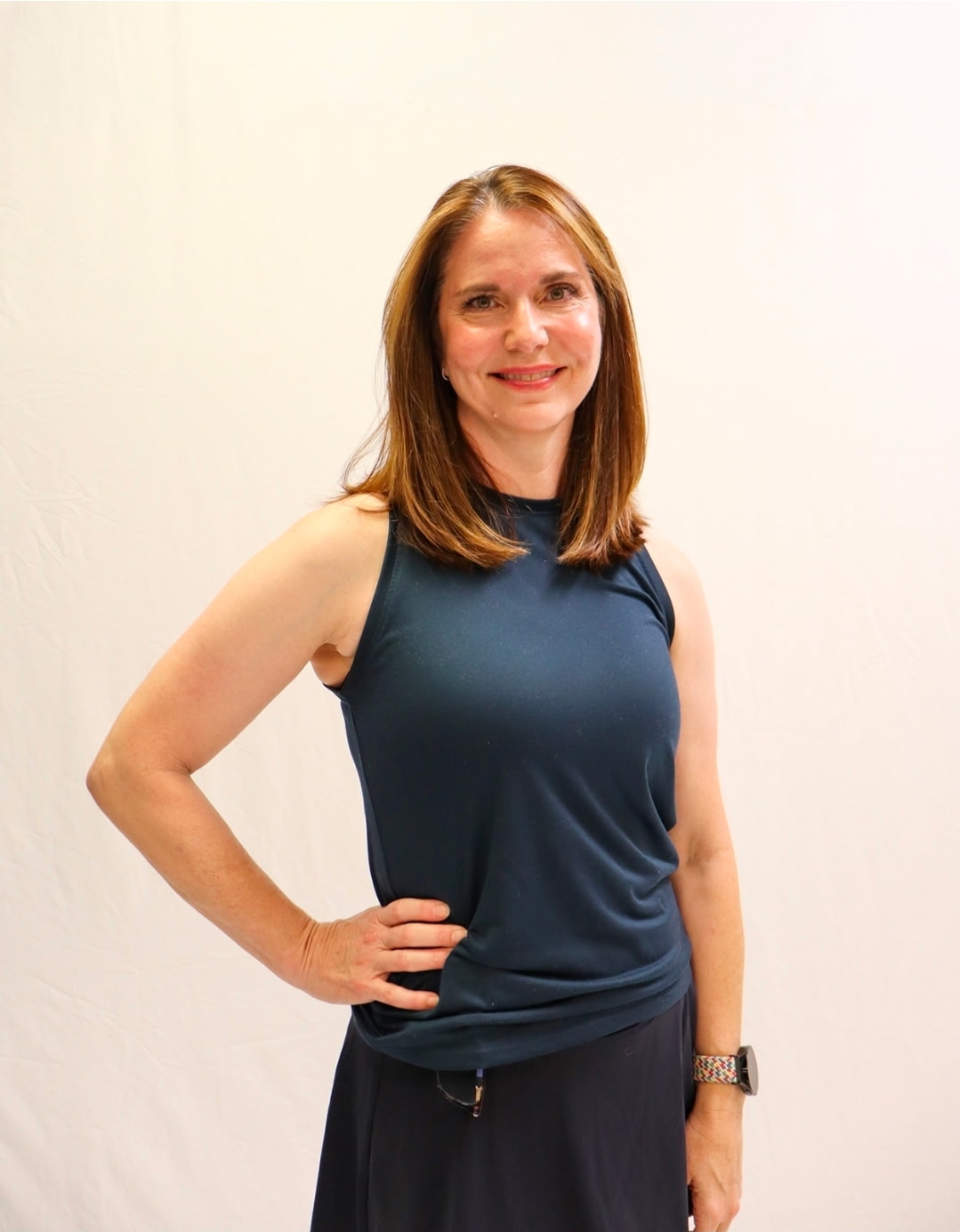 A woman with straight, shoulder-length brown hair smiles at the camera, standing against a plain white background. She wears a sleeveless dark top, a skirt, a watch, and has her right hand on her hip. -Showstoppers Dance Studio