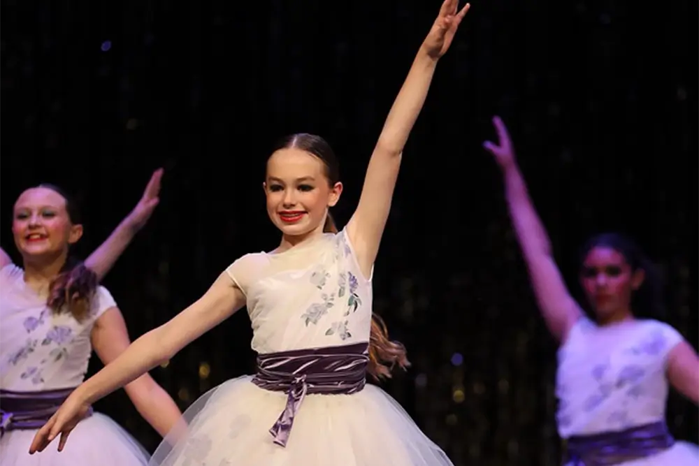 Three young dancers in white dresses with purple sashes perform on stage, smiling with one arm raised each. The background is dark with faint sparkles. -Showstoppers Dance Studio