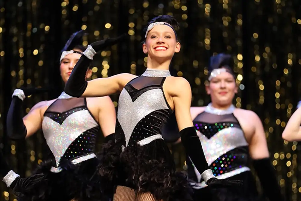 Three dancers in sparkling black and silver costumes perform onstage, smiling with gloved arms raised against a backdrop of golden lights. -Showstoppers Dance Studio