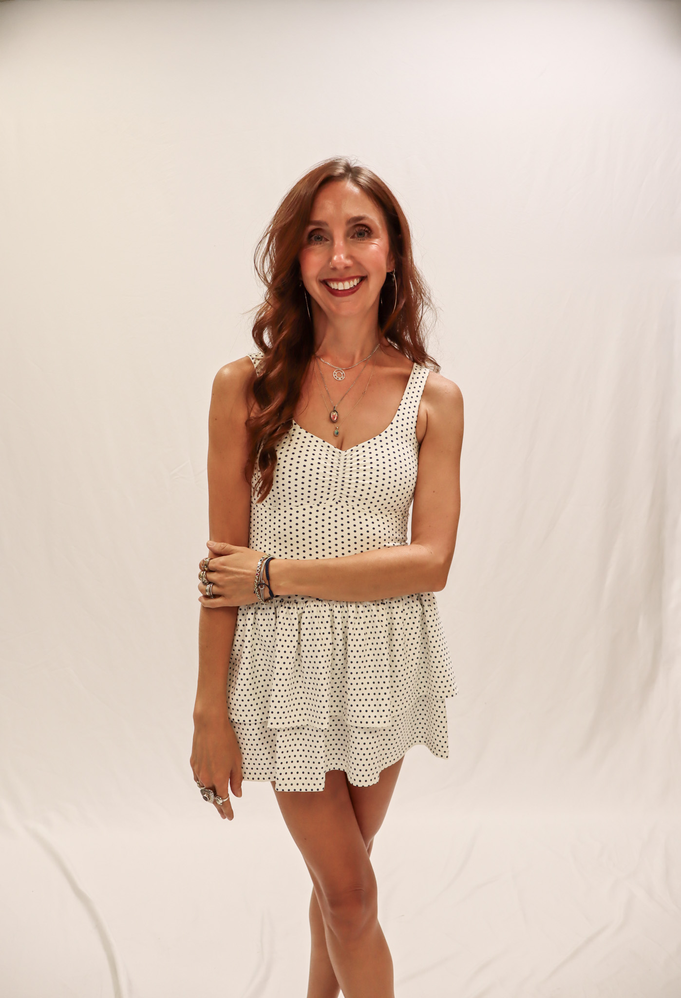 A woman in a white, sleeveless, polka-dot dress stands smiling against a plain white backdrop, with her arms relaxed and one hand holding her opposite arm. -Showstoppers Dance Studio