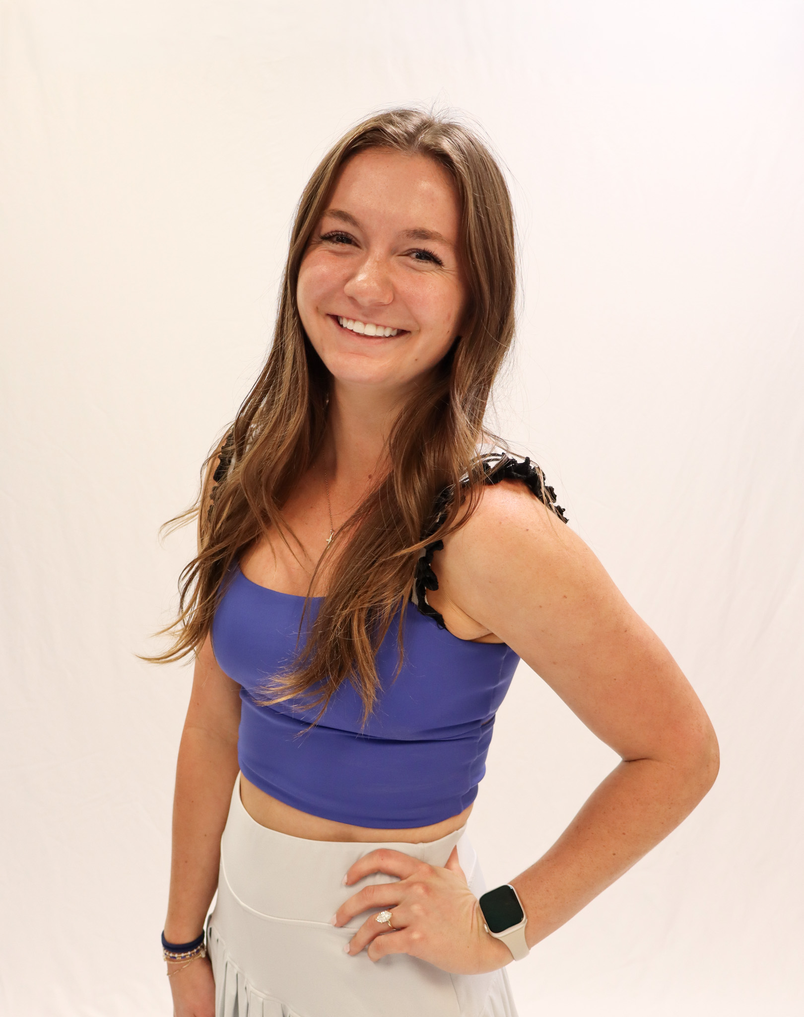Young woman with long brown hair smiles at the camera, wearing a purple crop top, light-colored skirt, and a smart watch, standing against a plain white background. -Showstoppers Dance Studio