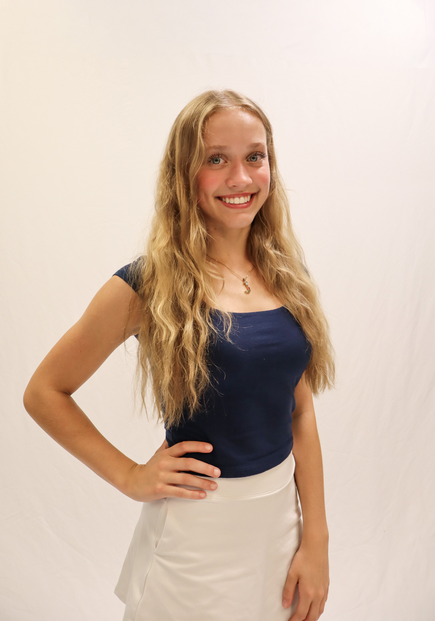 A young woman with long blonde hair wearing a navy blue top and white skirt smiles at the camera against a plain white background. -Showstoppers Dance Studio