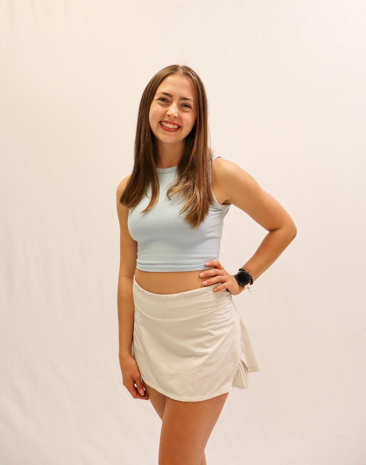 A young woman with long brown hair, wearing a light blue sleeveless crop top and a white skirt, smiles while standing against a plain white background. -Showstoppers Dance Studio