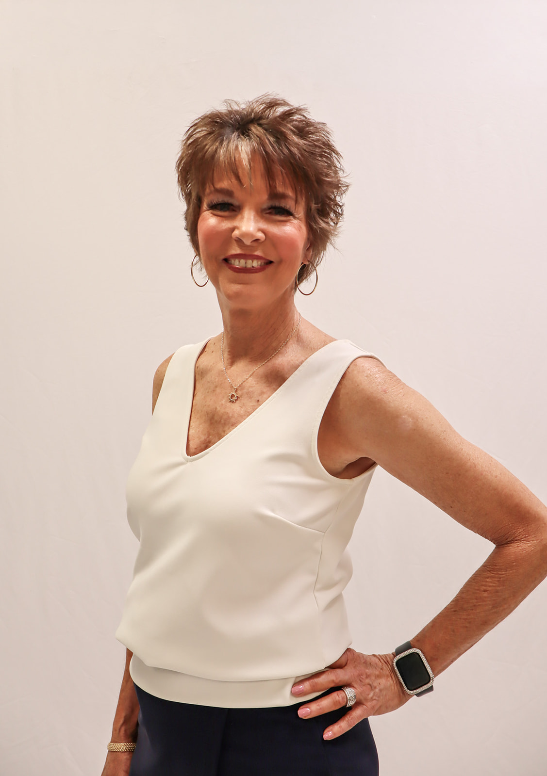 A smiling woman with short brown hair wearing a sleeveless white top, dark pants, hoop earrings, a necklace, a smart watch, and a ring stands against a plain light background with one hand on her hip. -Showstoppers Dance Studio