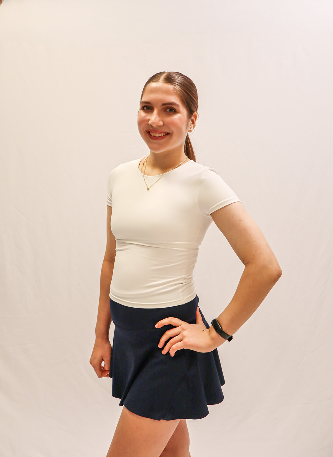 A young woman with long brown hair in a ponytail smiles, posing in a white fitted top and navy blue skirt against a plain white background. She has one hand on her hip and wears a necklace and a black wristband. -Showstoppers Dance Studio