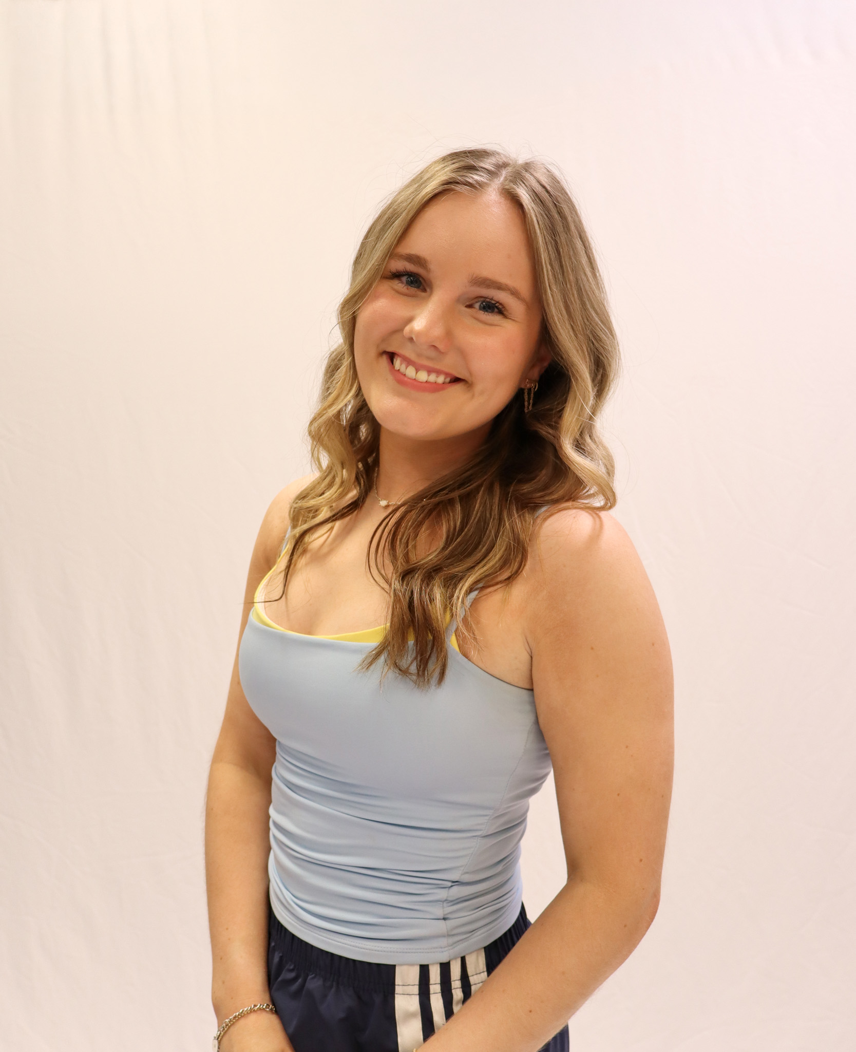 A young woman with long, wavy blonde hair smiles at the camera. She is wearing a light blue tank top and navy blue pants. The background is plain white. -Showstoppers Dance Studio