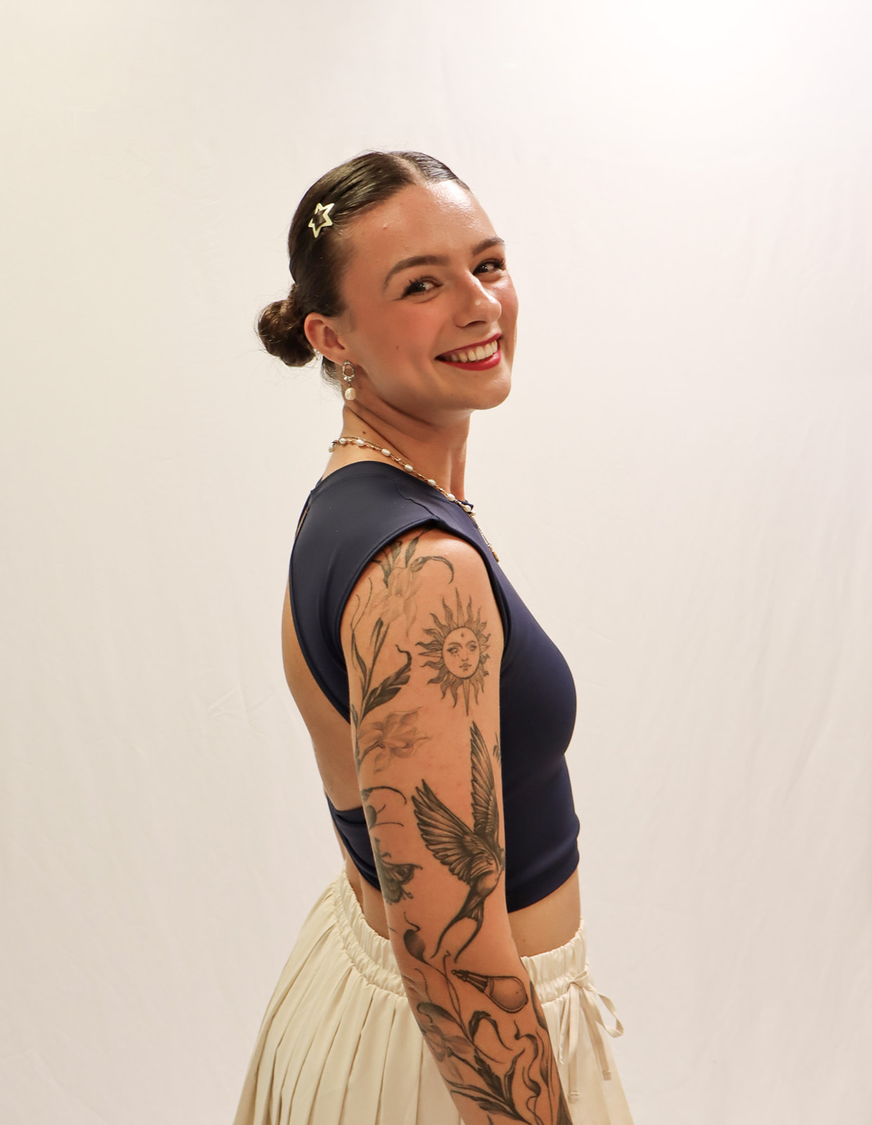 A smiling person with hair in buns, wearing a navy sleeveless top and cream skirt, showing tattoos of a bird, sun, and flowers on their arm, stands against a plain white background. -Showstoppers Dance Studio