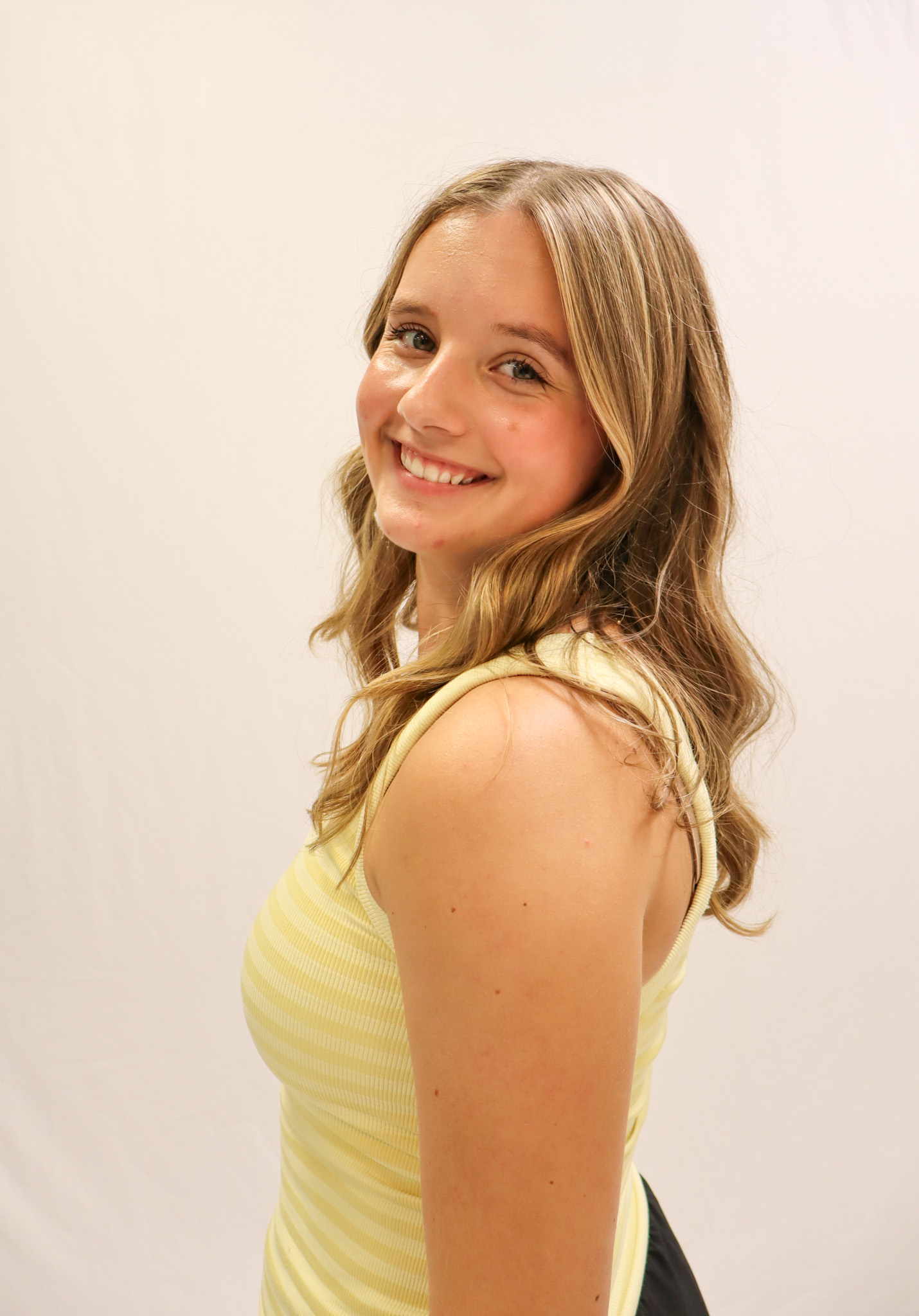 A young woman with wavy blonde hair, wearing a sleeveless yellow striped top, smiles while looking over her shoulder against a plain white background. -Showstoppers Dance Studio
