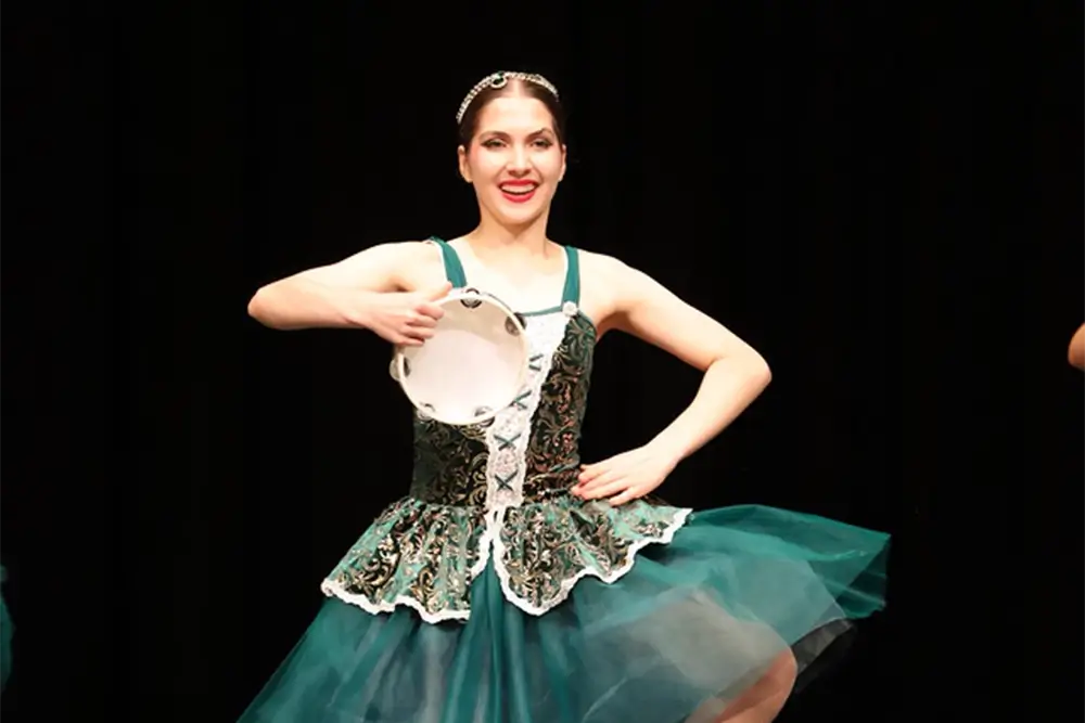 A ballerina in a green and white tutu dances on stage, smiling and holding a tambourine. The background is black, making her stand out. -Showstoppers Dance Studio