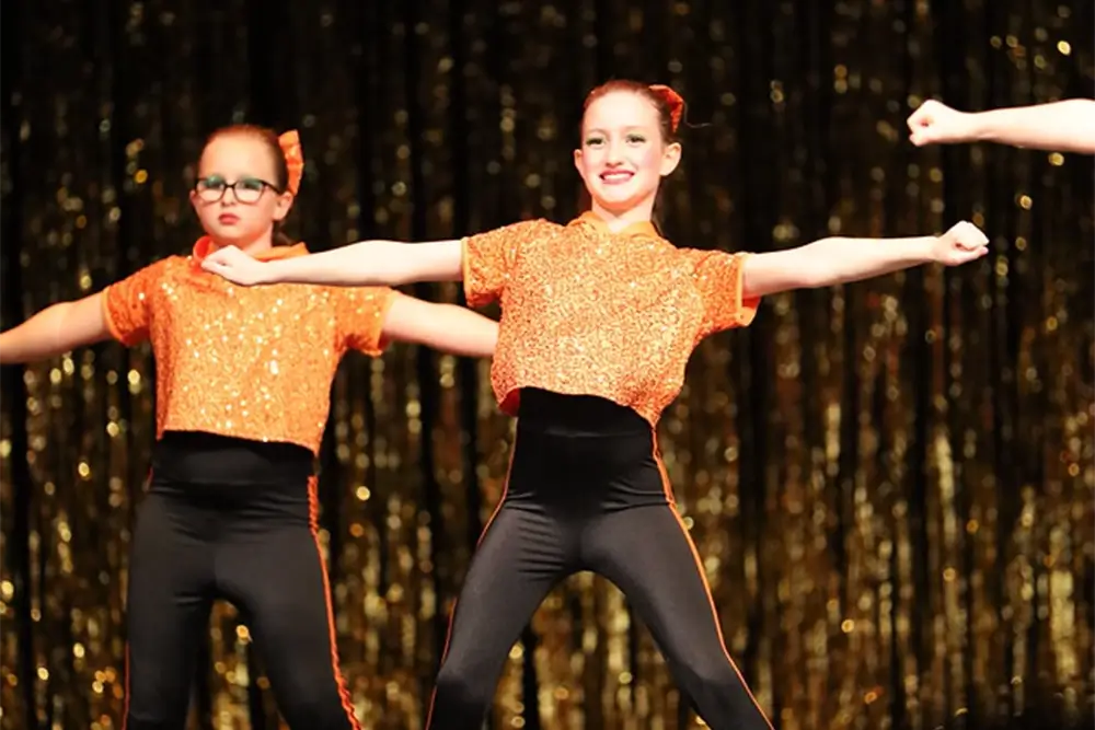 Two young dancers in orange sequined tops and black pants perform on stage with arms extended. The background is decorated with gold streamers. -Showstoppers Dance Studio