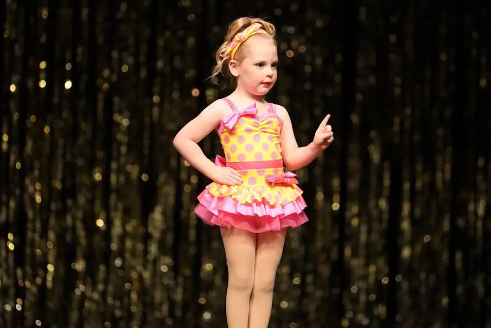 A young girl in a pink and yellow polka dot dance costume stands on stage with one hand on her hip and the other pointing up, against a glittery black and gold backdrop. -Showstoppers Dance Studio