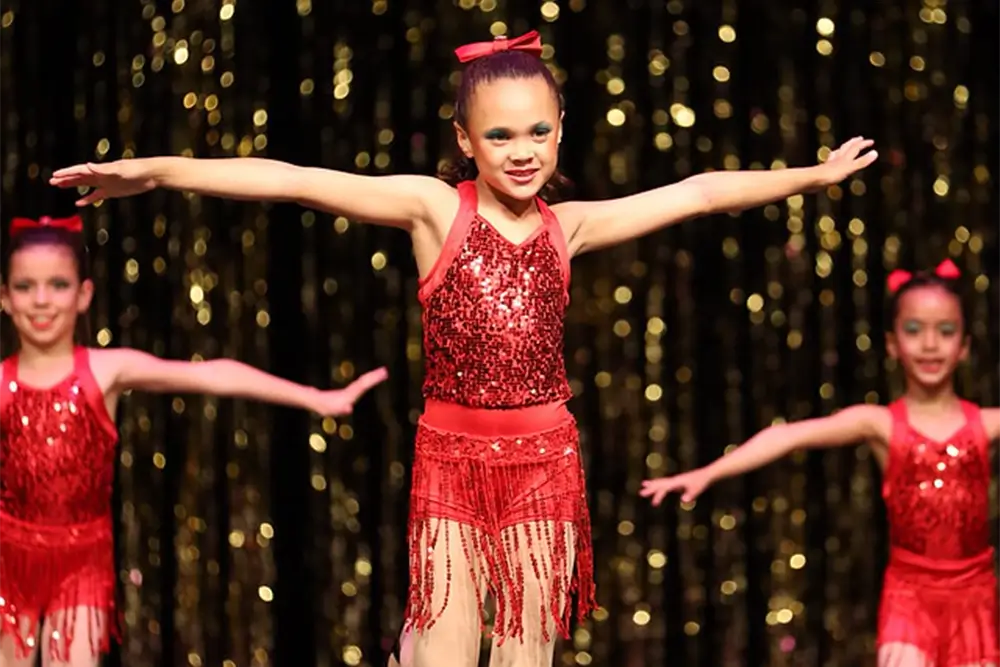 Three young girls in red sequined dance costumes with fringe and bows perform with arms outstretched on stage, in front of a shiny gold backdrop. -Showstoppers Dance Studio