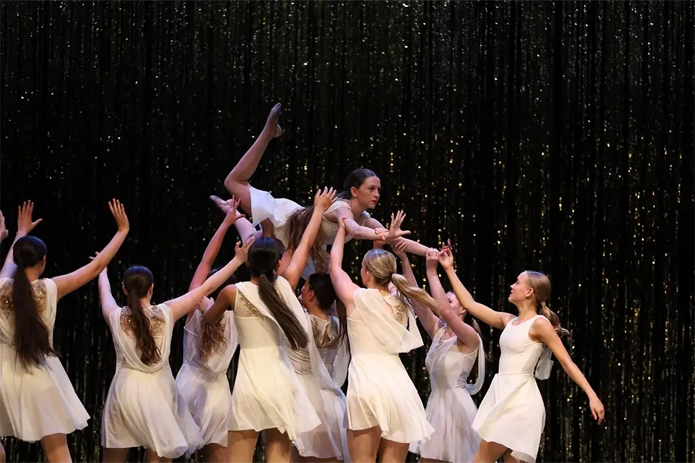 A group of dancers in white dresses lift a teammate above their heads on stage, with black and gold shimmering curtains in the background. -Showstoppers Dance Studio