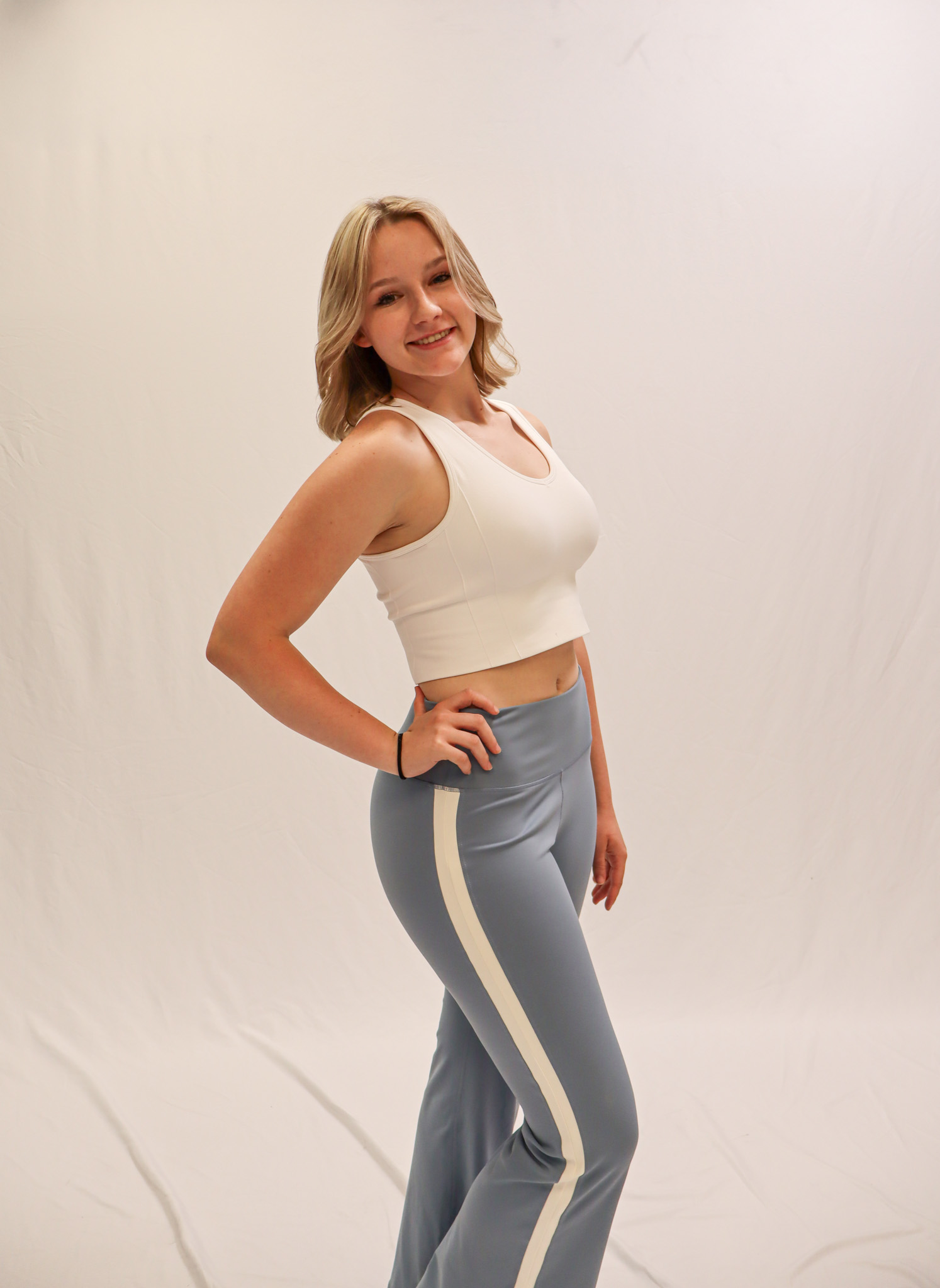 A young woman with blonde hair stands smiling, wearing a white crop top and light blue pants with white stripes, posing against a plain white background. -Showstoppers Dance Studio
