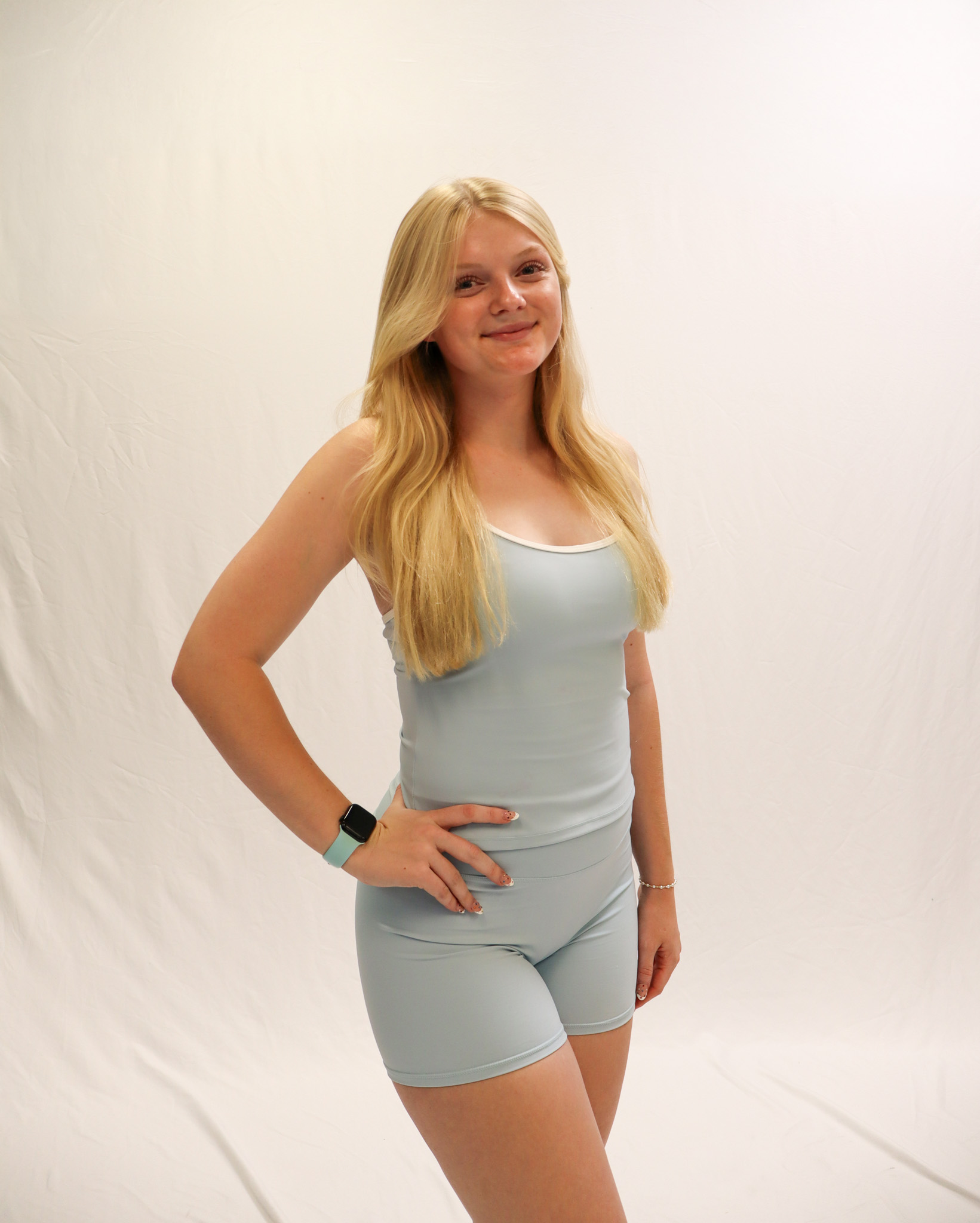 A young woman with long blonde hair wears a light blue tank top and matching shorts, standing against a plain white background and smiling at the camera. -Showstoppers Dance Studio