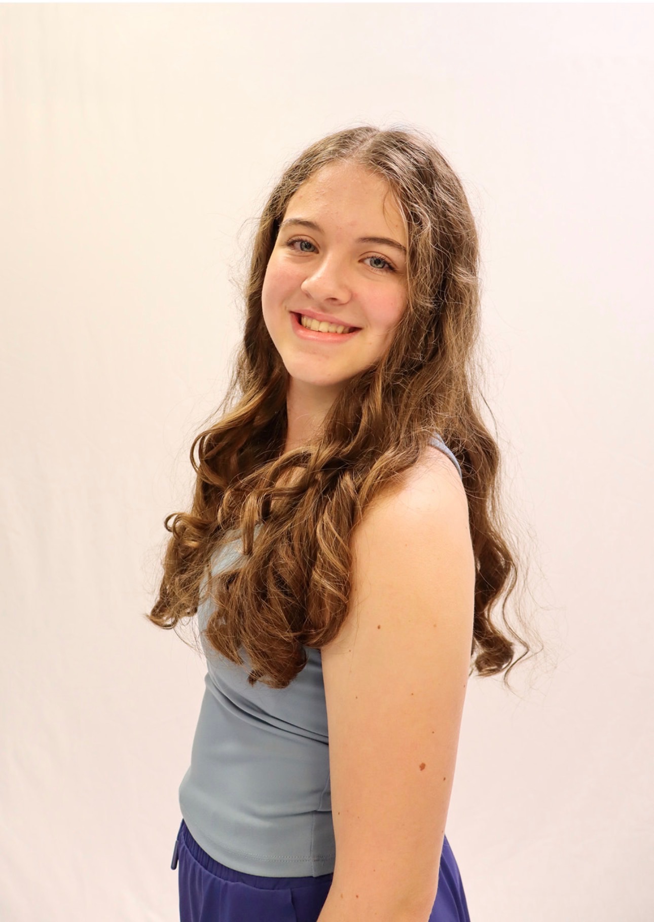 A young woman with long, curly brown hair smiles at the camera. She is wearing a light blue sleeveless top and dark blue pants, standing in front of a plain white background. -Showstoppers Dance Studio