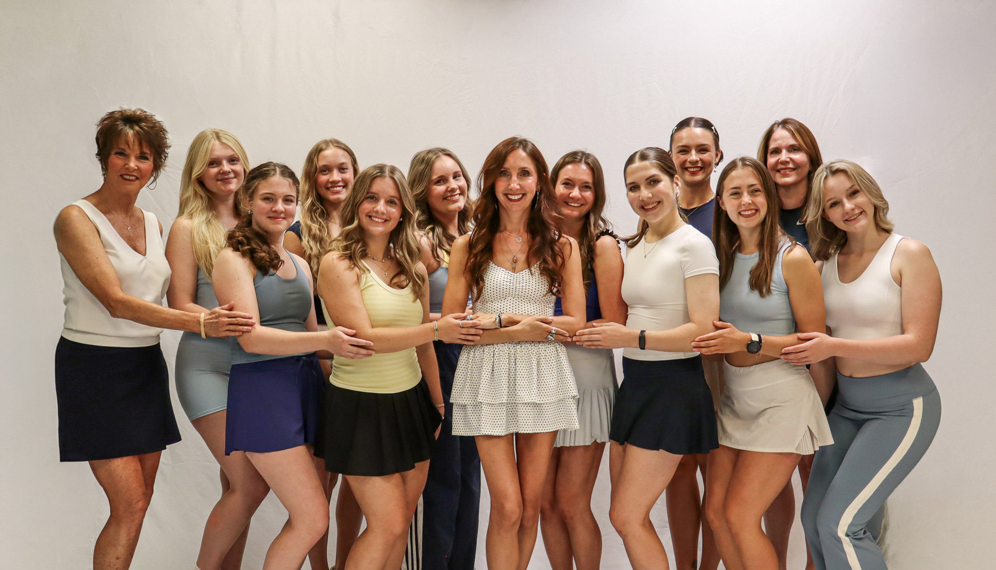 Fourteen women stand together in a row against a white background, smiling and holding hands. Most wear athletic or casual outfits in light colors. They appear happy and friendly. -Showstoppers Dance Studio