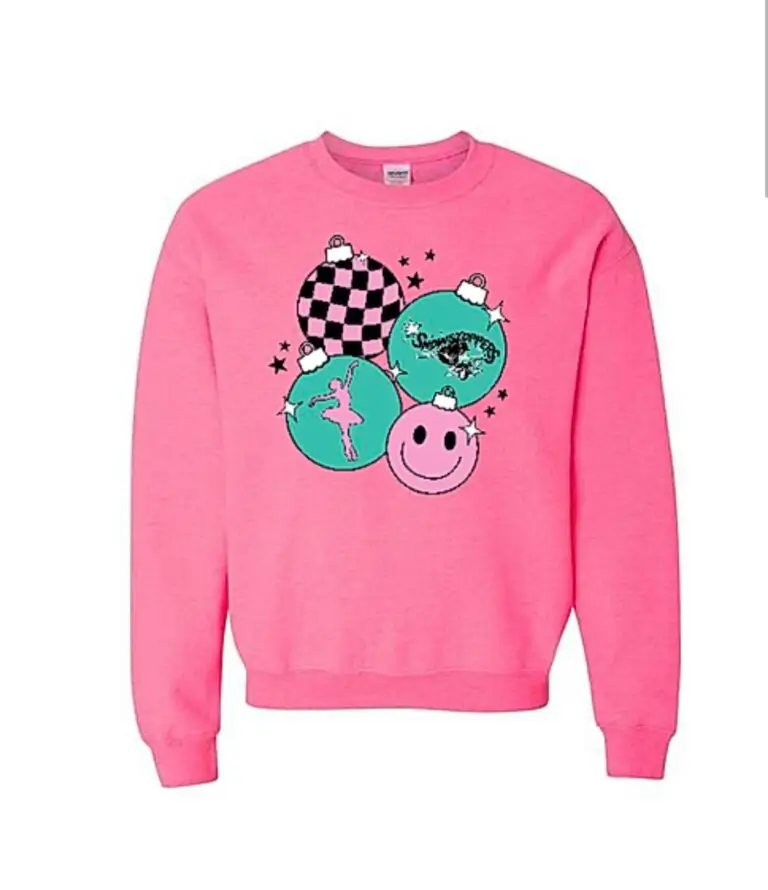 A pink sweatshirt features three teal Christmas ornaments with different designs: a checkerboard pattern, a ballerina, and a winter scene. Below them is a large pink smiley face and small black stars scattered around. -Showstoppers Dance Studio