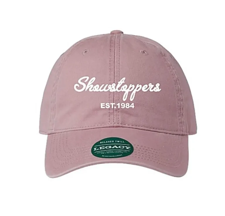 A mauve baseball cap with white text reading Showstoppers EST. 1984 on the front and a green Legacy sticker on the brim. -Showstoppers Dance Studio