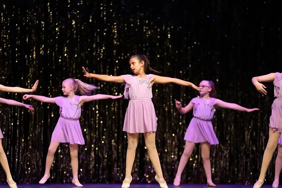 Five young girls wearing matching lilac dresses perform a dance routine on stage, standing in formation with arms extended. The background is decorated with shimmering black and gold tinsel. -Showstoppers Dance Studio