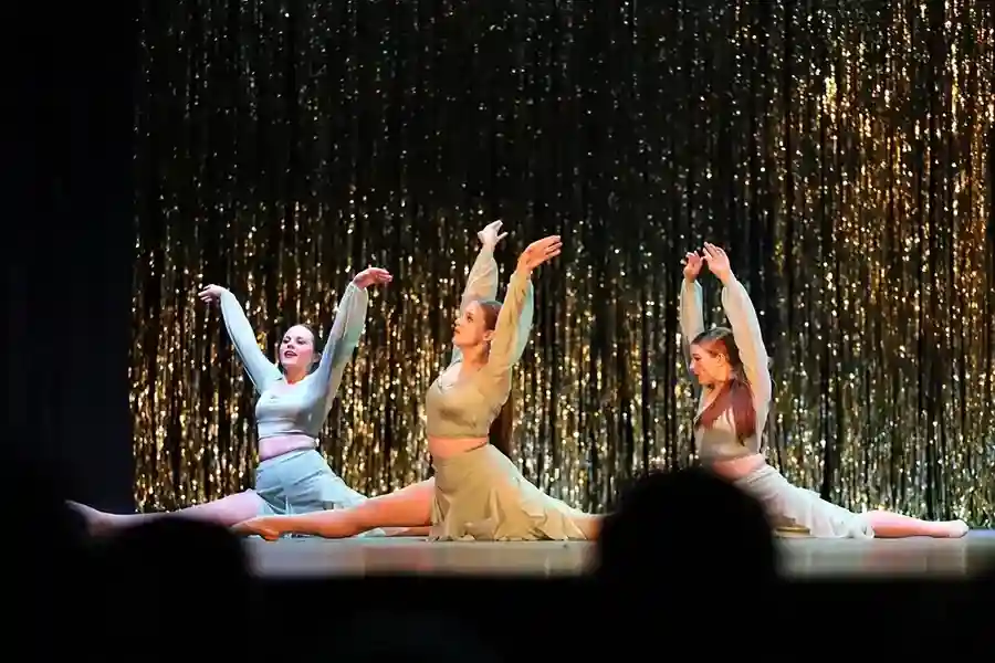 Three dancers in matching outfits perform a split with arms raised on stage, set against a backdrop of shimmering gold streamers. -Showstoppers Dance Studio