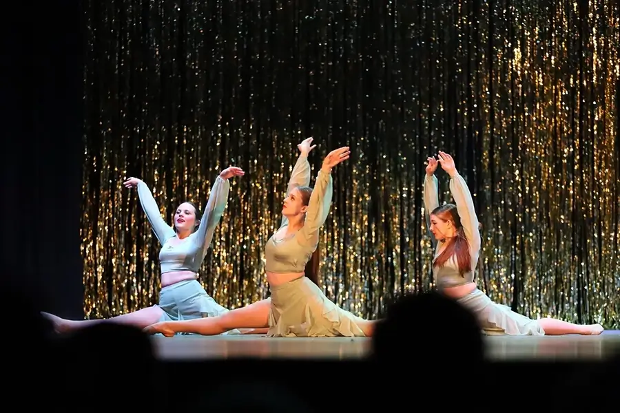 Three dancers in light green costumes perform splits on stage with arms raised, in front of shimmering gold curtains. The scene is brightly lit. -Showstoppers Dance Studio