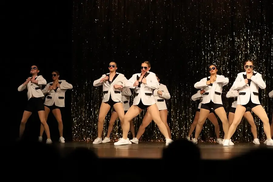 A group of dancers in white blazers, black shorts, and sunglasses perform energetically on stage in front of a glittering backdrop. -Showstoppers Dance Studio