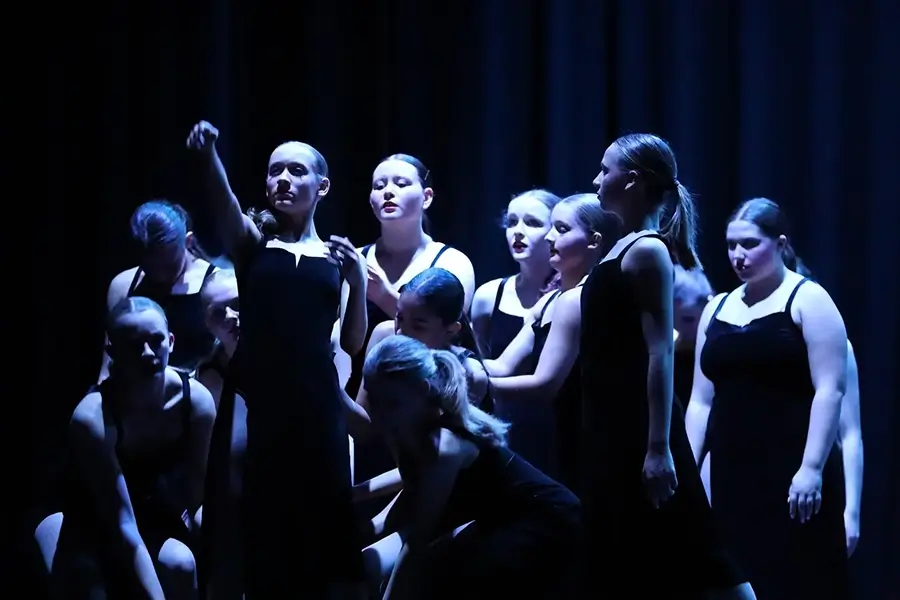 A group of dancers in black dresses perform on stage under dramatic blue lighting, with one dancer extending her arm forward and others clustered around her. -Showstoppers Dance Studio