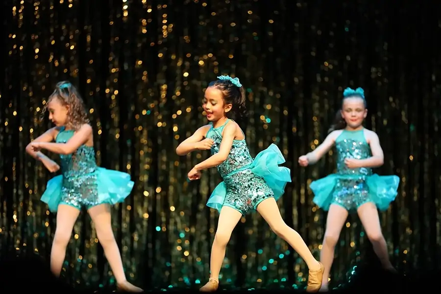 Three young girls in sparkly turquoise dance costumes perform on stage, dancing energetically in front of a gold tinsel curtain backdrop. -Showstoppers Dance Studio